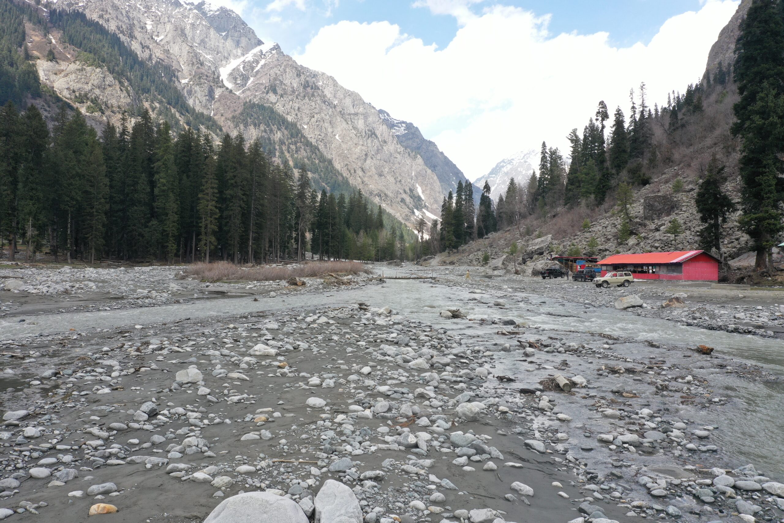 A wide, rocky riverbed in a mountainous valley with pine trees and a distant red building, representing the stunning scenery available with Kumrat Valley Glamping Deals.