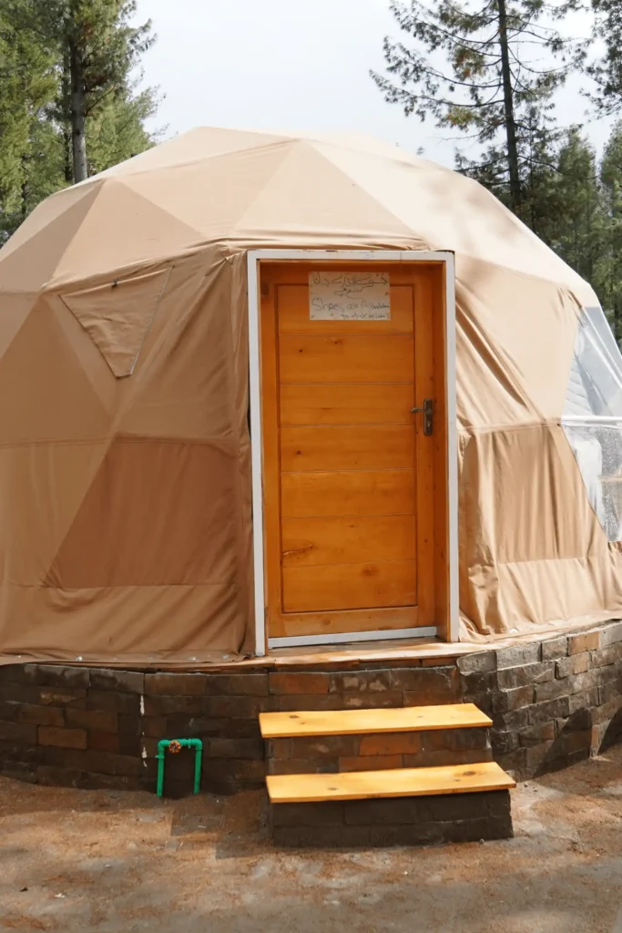 A luxurious glamping dome, perfect for glamping in Pakistan, with a wooden door and steps set against a natural, treed backdrop.