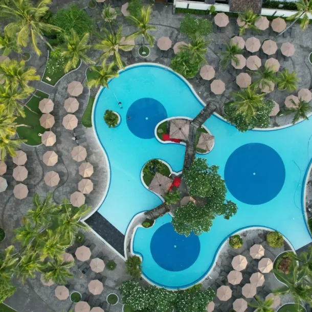 An aerial view of a large, multi-sectioned swimming pool surrounded by numerous lounge chairs and palm trees, clearly illustrating how resorts are different from hotels.