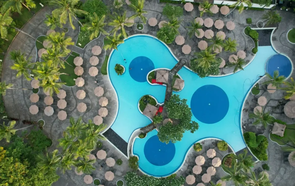 An aerial view of a large, multi-sectioned swimming pool surrounded by numerous lounge chairs and palm trees, clearly illustrating how resorts are different from hotels.