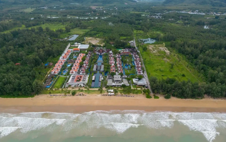 An aerial view of a large beach resort complex with a swimming pool and buildings, juxtaposed with a mountain range in the background, perfectly illustrating the choice between Beach Resorts vs Mountain Resorts.