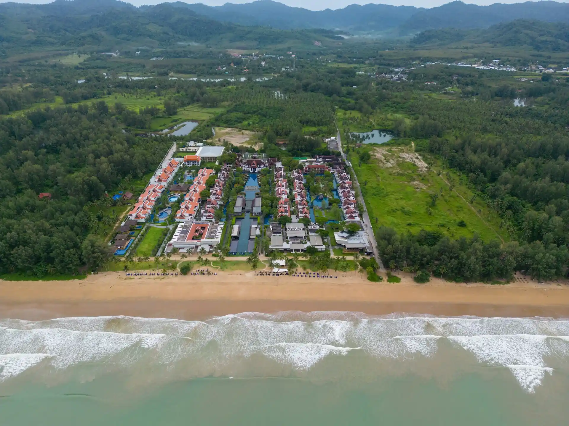 An aerial view of a large beach resort complex with a swimming pool and buildings, juxtaposed with a mountain range in the background, perfectly illustrating the choice between Beach Resorts vs Mountain Resorts.